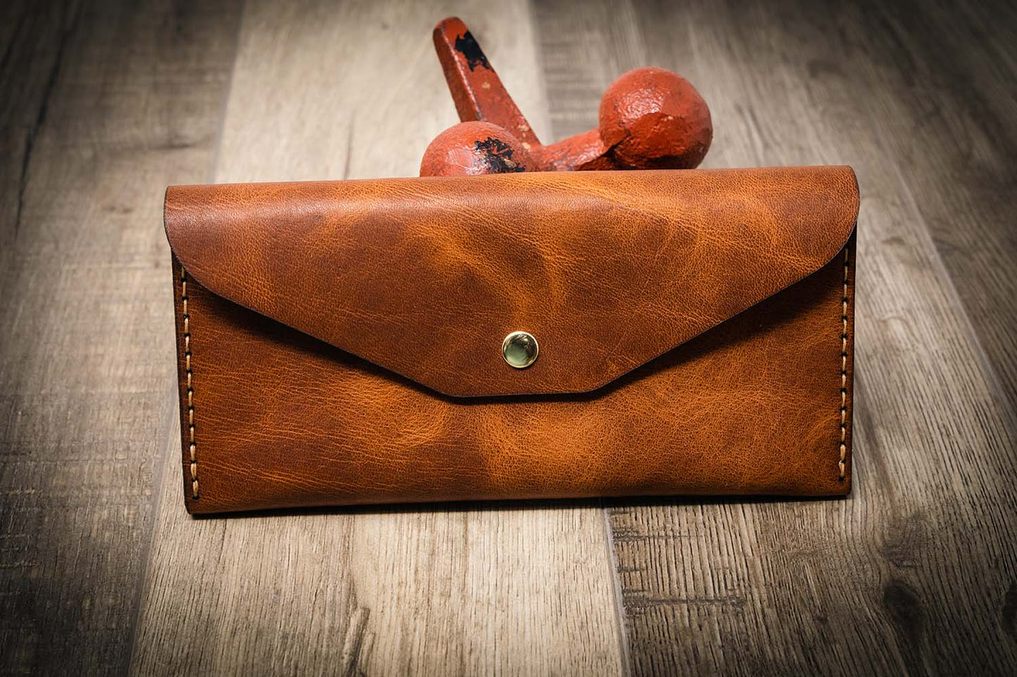 Women's leather wallet clutch in chestnut brown, handmade in the USA, displayed on a wooden surface with a rustic metal tool in the background.
