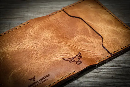 Close-up of a handcrafted leather wallet in a warm tan color, featuring visible natural grain patterns, contrast stitching, and an embossed bull head logo from Bull Sheath Leather.