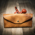 Handmade women's long wallet in saddle tan leather, featuring a brass button, on a wooden surface with a rustic red weight in the background.

