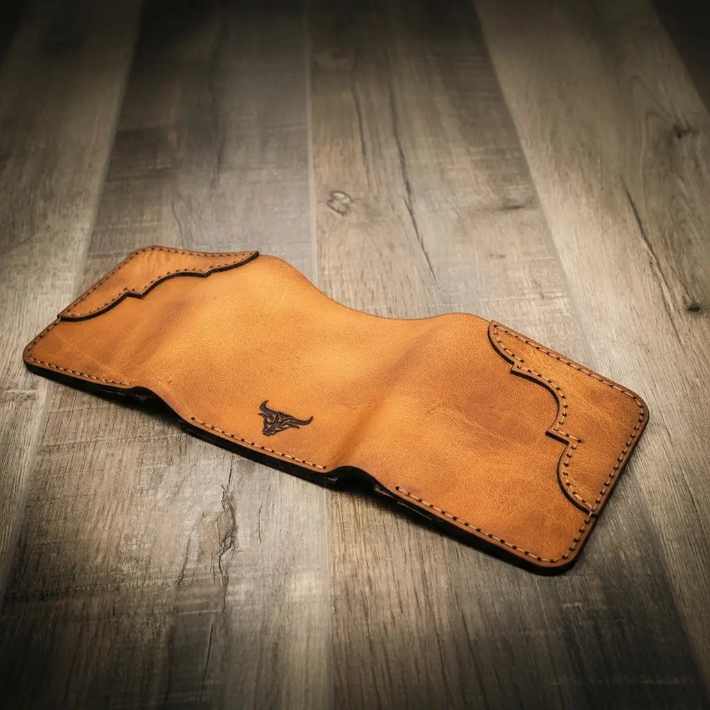 Trinity western trifold wallet in saddle tan leather, featuring intricate stitching and a bull logo, displayed open on a wooden surface.
