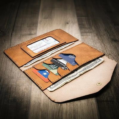 Handmade women's long wallet in saddle tan leather, open to show card slots and ID window, on a wooden surface.
