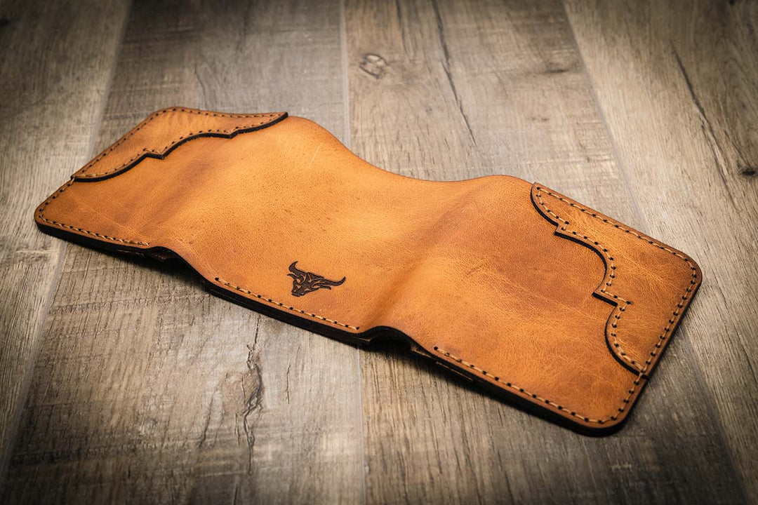 Trinity western trifold wallet in saddle tan leather, featuring intricate stitching and a bull logo, displayed open on a wooden surface.