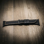 Apple Watch Leather Band in black full-grain leather with tan stitching on wood floor.