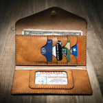 Handmade women's long wallet in saddle tan leather, open to show card slots, ID window, and cash compartment, on a wooden surface.
