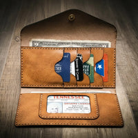 Handmade women's long wallet in saddle tan leather, open to show card slots, ID window, and cash compartment, on a wooden surface.
