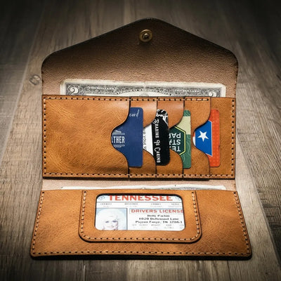 Handmade women's long wallet in saddle tan leather, open to show card slots, ID window, and cash compartment, on a wooden surface.
