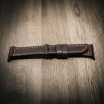 Brown full-grain leather Apple Watch band with tan stitching and black buckle on wooden surface