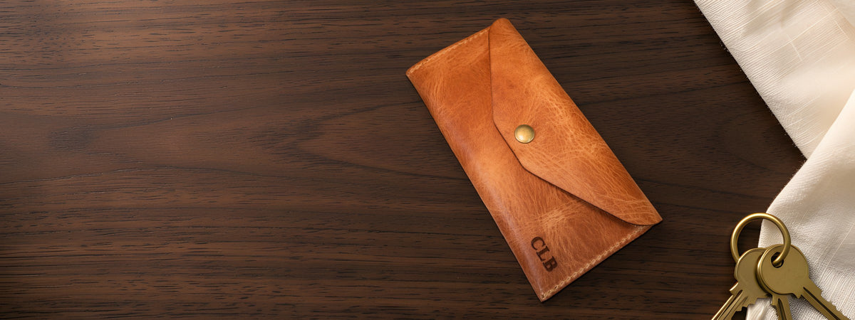 Badge Clutch Wallet in chestnut full-grain leather closed on a dark walnut table beside keys and a linen cloth