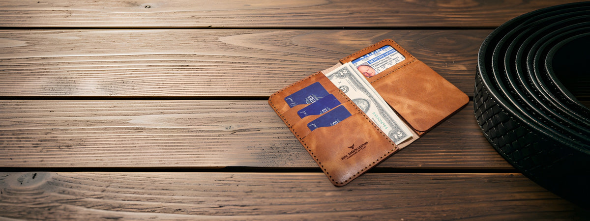 Badge Long Wallet in chestnut full-grain leather open with cards and cash on dark wood planks beside a coiled black belt