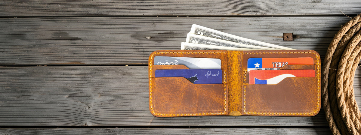 Rio Grande Bifold Wallet in chestnut full-grain leather, open with cards and cash, lying on weathered grey barnwood beside a coiled lariat rope