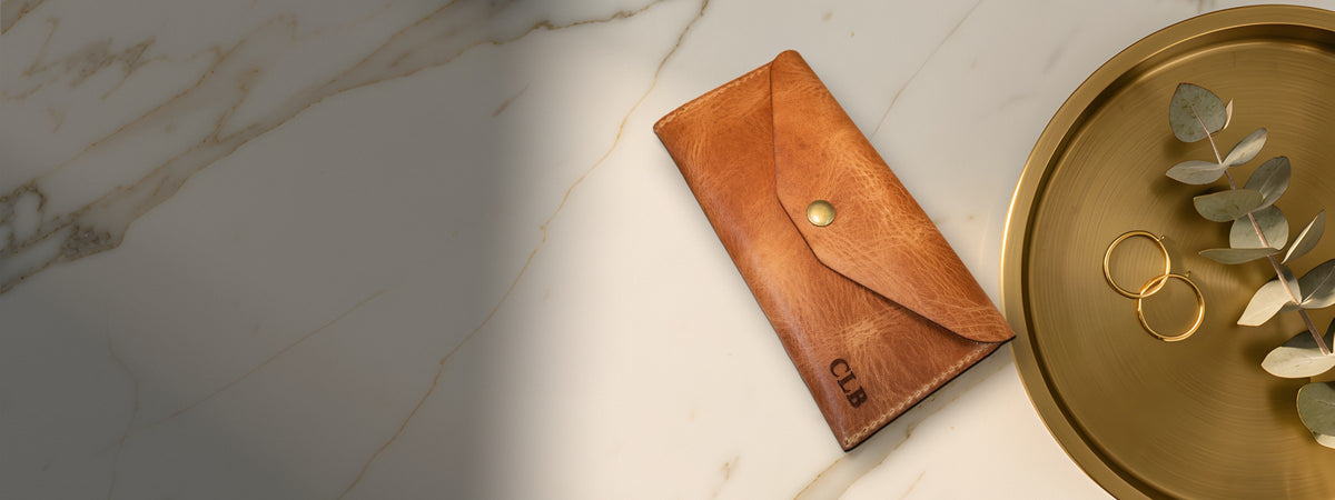 Sugar Land Clutch Wallet in chestnut full-grain leather closed on white marble beside a brass tray