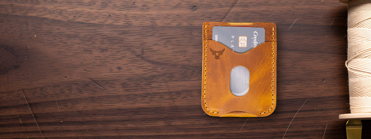 The Keller Money Clip in tan full-grain leather, card-slot front with the bull logo, holding a card on a dark walnut table beside a spool of thread