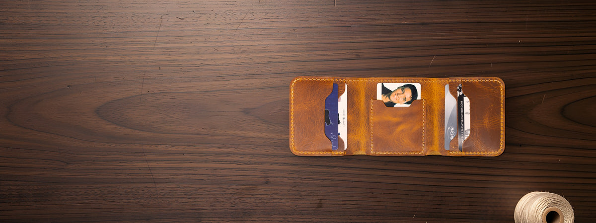 The Trinity Trifold Wallet in chestnut full-grain leather open with cards on a dark walnut table beside a spool of thread