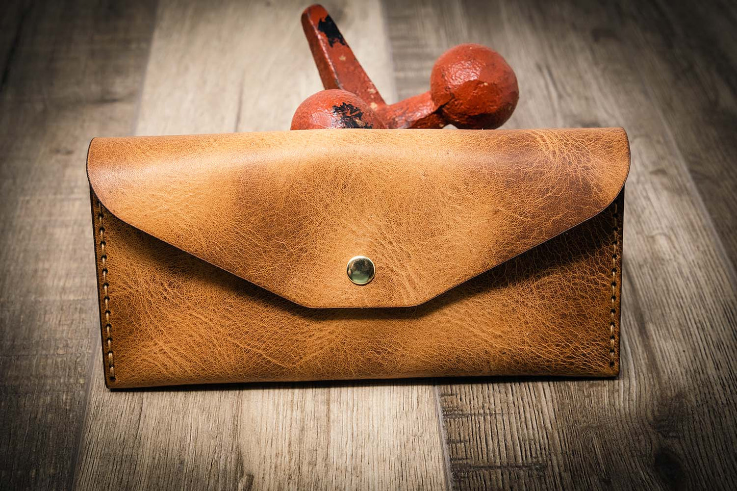 Handmade women's long wallet in saddle tan leather, featuring a brass button, on a wooden surface with a rustic red weight in the background.